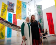 Two women smiling for the camera in front of a large gallery room full of long rectangular textile pieces of red, yellow and blue