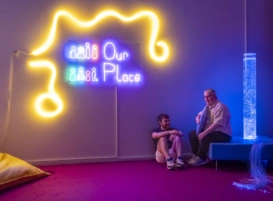 Two men sit under a lit-up sign that reads Our Place. One man sits on the floor, the other sits on a bench holding a cushion. Behind him is a bubble tube.