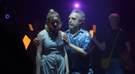 A man is looking at a younger woman holding her shoulders. They are in a dark room with orange rectangular lights behind them.