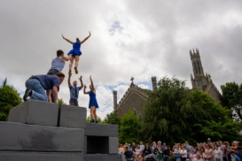 Five people on top of grey blocks, one woman is jumping into the arms of a man and a woman who have their arms outstretched. To the right is a view of Carlow Cathedral behind trees and a large audience watching from below.