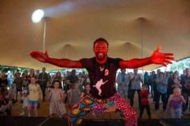 A man looking into the camera with his arms outstretched, behind him is a room full of people facing him, ready to dance