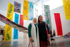 Two women smiling for the camera in front of a large gallery room full of long rectangular textile pieces of red, yellow and blue