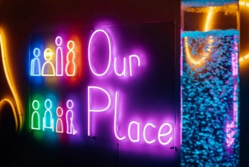 Black background with a Purple neon sign spelling out 'Our Place', alongside neon people figures, in foreground is a blue glitter lamp