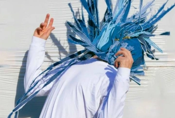 Artist Barry Fitzgerald promotional imagery for Mourning is a Muscle. Fitzgerald stands against a white wall wearing a white button down shirt with his arms raised and his head back. On his head, he is wearing a blue denim tassel mask is moving in different directions.