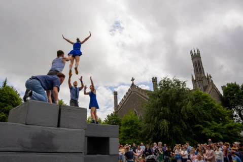 Five people on top of grey blocks, one woman is jumping into the arms of a man and a woman who have their arms outstretched. To the right is a view of Carlow Cathedral behind trees and a large audience watching from below.