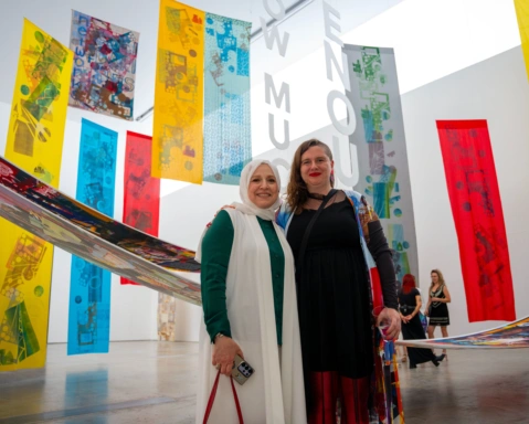 Two women smiling for the camera in front of a large gallery room full of long rectangular textile pieces of red, yellow and blue