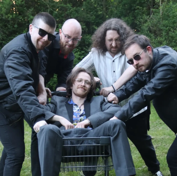 Five male band members pose, one sits in a shopping trolley in the middle and the other four stand behind him