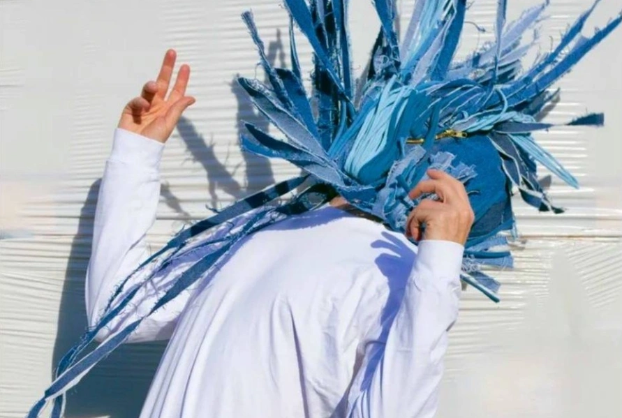 Artist Barry Fitzgerald promotional imagery for Mourning is a Muscle. Fitzgerald stands against a white wall wearing a white button down shirt with his arms raised and his head back. On his head, he is wearing a blue denim tassel mask is moving in different directions.